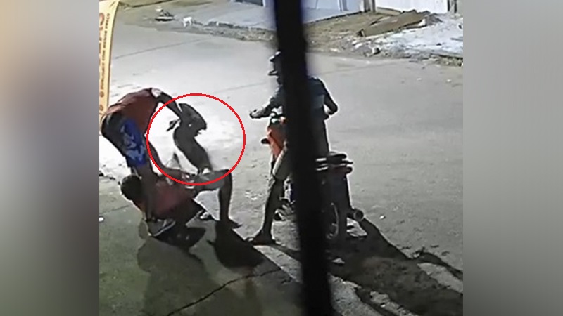 Homem agredindo outro no chão com objeto, enquanto um motociclista observa a cena.