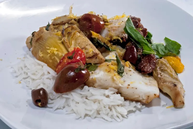 Prato de peixe branco com arroz, alcachofras, tomates e azeitonas em um prato branco.