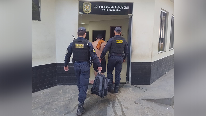 Dois guardas municipais escoltando um homem algemado para dentro de uma delegacia.