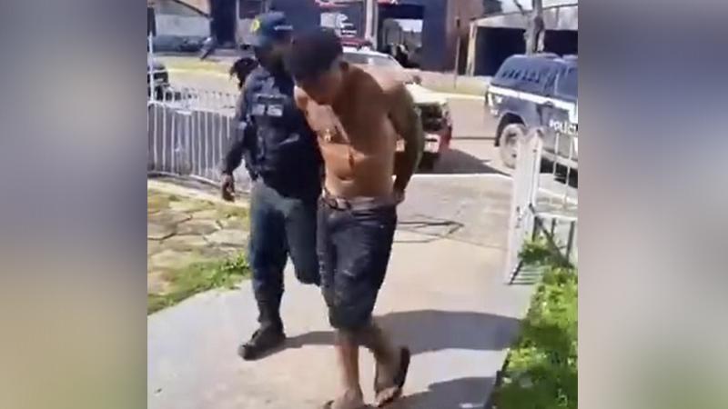 Policial fardado escoltando um homem sem camisa em uma rua durante o dia.