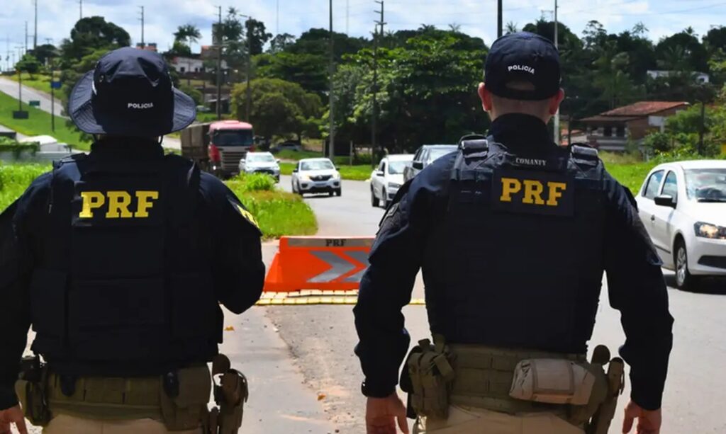 Dois policiais da PRF em um posto de fiscalização em uma estrada, com carros ao fundo.