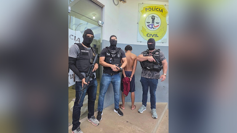 Três policiais civis mascarados com coletes à prova de balas e armas, ao lado de um homem detido de costas, em frente a uma delegacia.