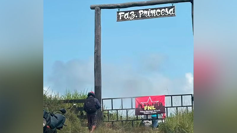 Portão de madeira da "Faz. Princesa" com faixa da FNL e uma pessoa caminhando.