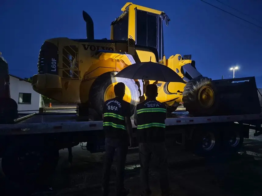 Fiscais da SEFA sob guarda-chuva observando pá carregadeira Volvo L60F sendo transportada à noite.
