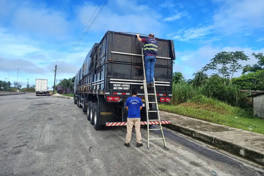 Dois fiscais da SEFA inspecionam a carga de um caminhão preto, um na escada e outro no chão.