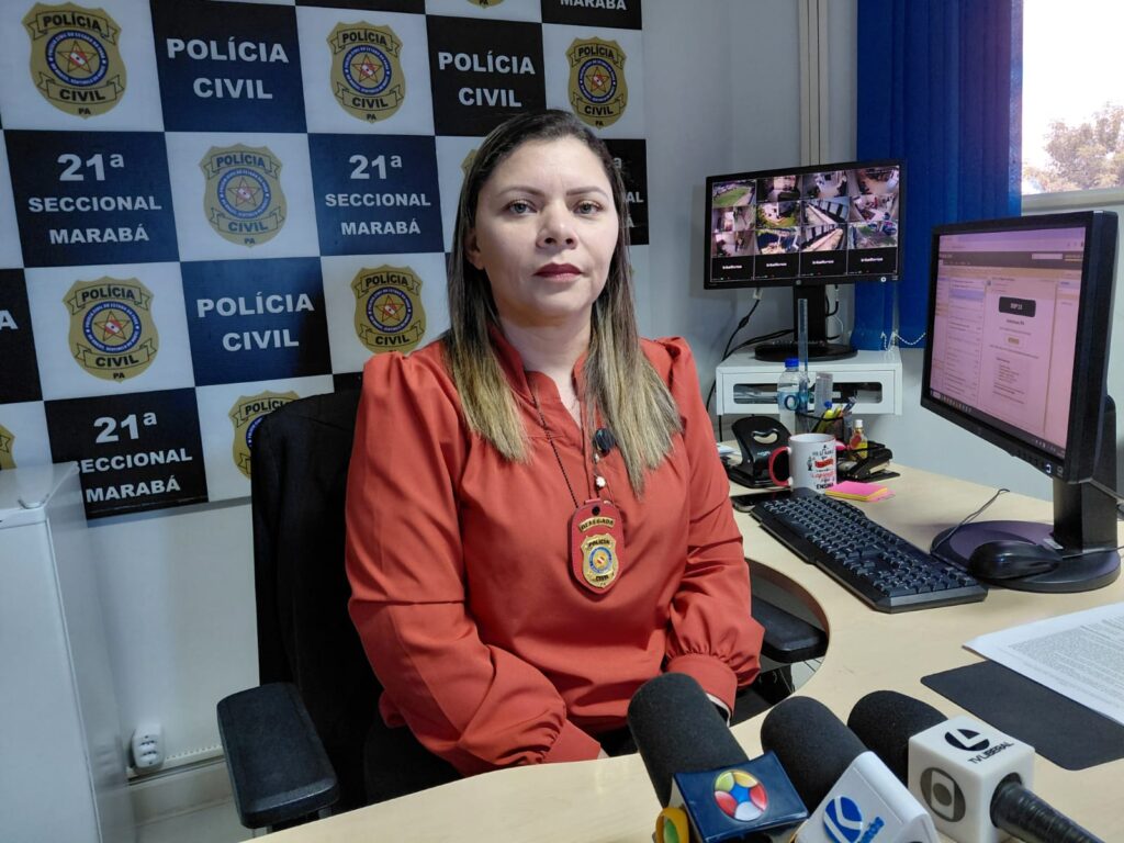 Mulher com blusa laranja e distintivo da Polícia Civil sentada em mesa de escritório.