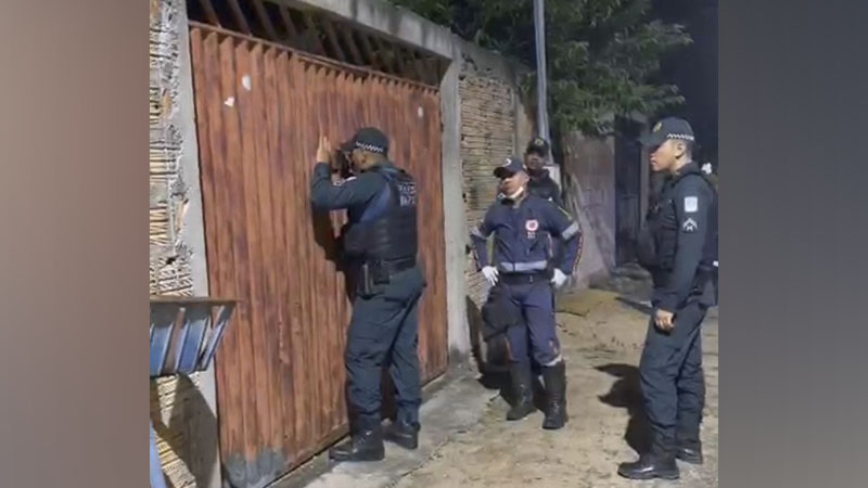 Policiais em uniforme escuro em frente a um portão de metal enferrujado, um deles batendo.