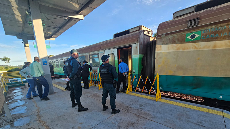 Trem da Estrada de Ferro Carajás parado em plataforma com policiais e civis, e minério vazado nos trilhos.
