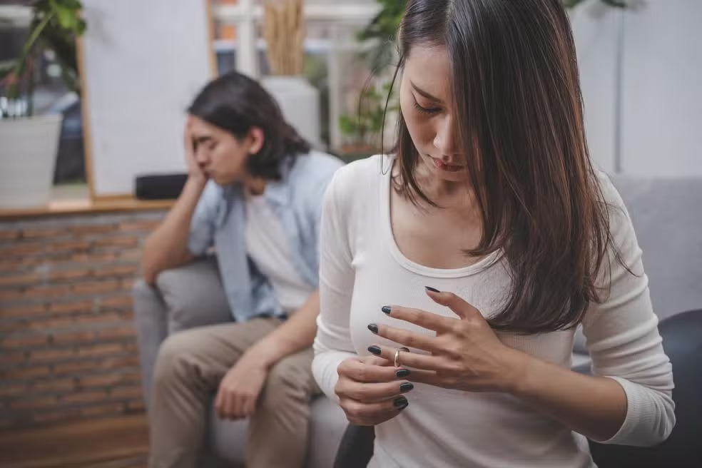 Mulher segurando um anel de casamento com homem triste ao fundo, sugerindo divórcio ou separação.