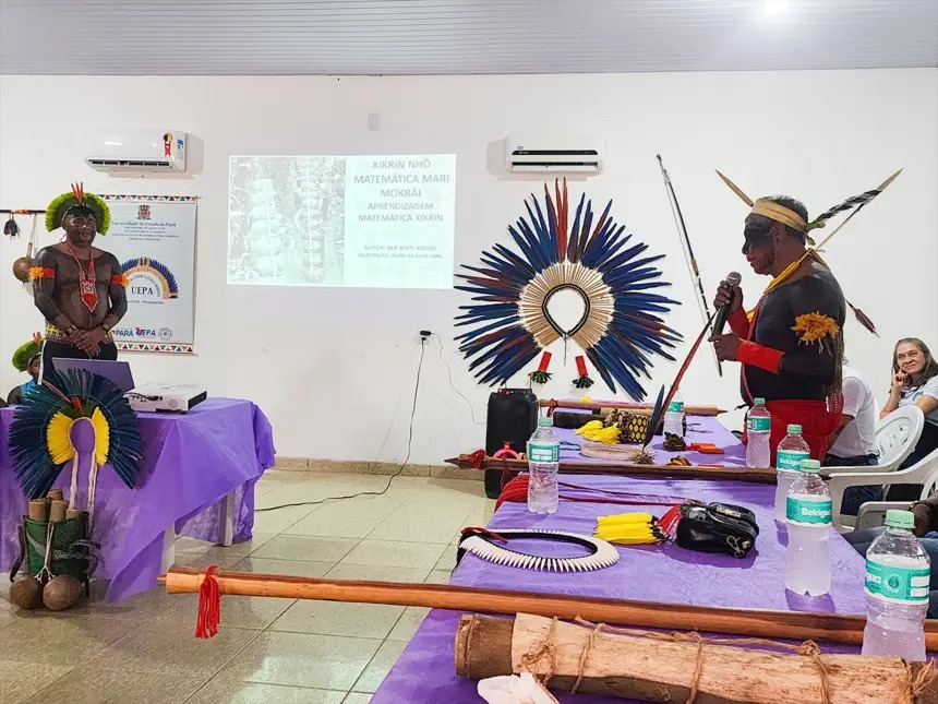 Homem indígena Xikrin com vestimentas tradicionais, falando ao microfone em evento educacional com artefatos culturais e projeção de slide.