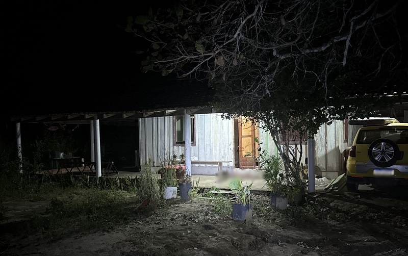 Imagem noturna de uma casa simples com um corpo no chão em frente à entrada.