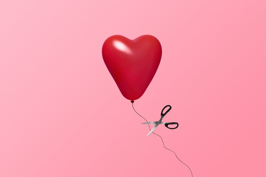 Balão vermelho em forma de coração com tesoura cortando a linha, sobre fundo rosa.