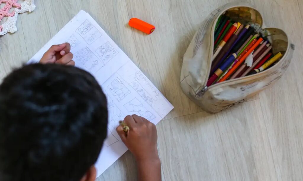 Criança desenhando em papel com lápis, ao lado de estojo com lápis de cor.