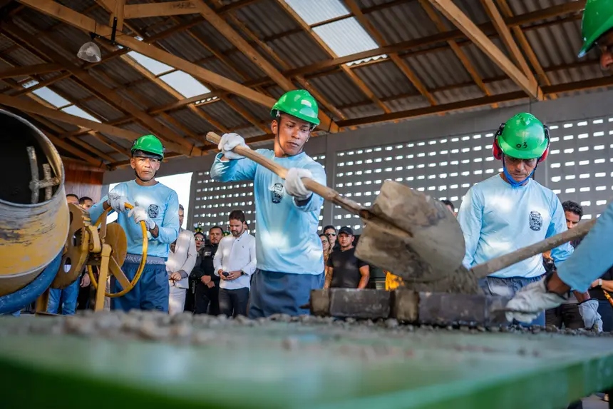 Homens com capacetes verdes trabalham com pá e cimento em ambiente de treinamento.