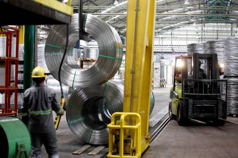 Trabalhador com capacete amarelo manuseando grandes bobinas de metal em um galpão industrial.
