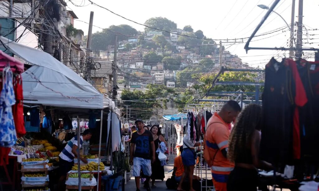 Feira de rua movimentada com pessoas e barracas de alimentos e roupas, e uma comunidade em um morro ao fundo.