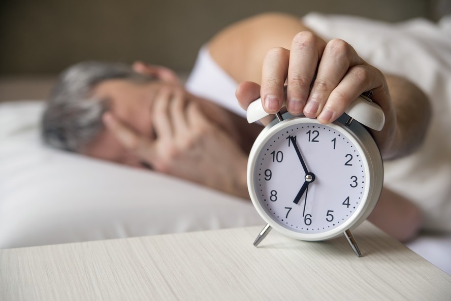 Pessoa na cama com as mãos no rosto, um despertador branco é segurado em primeiro plano.