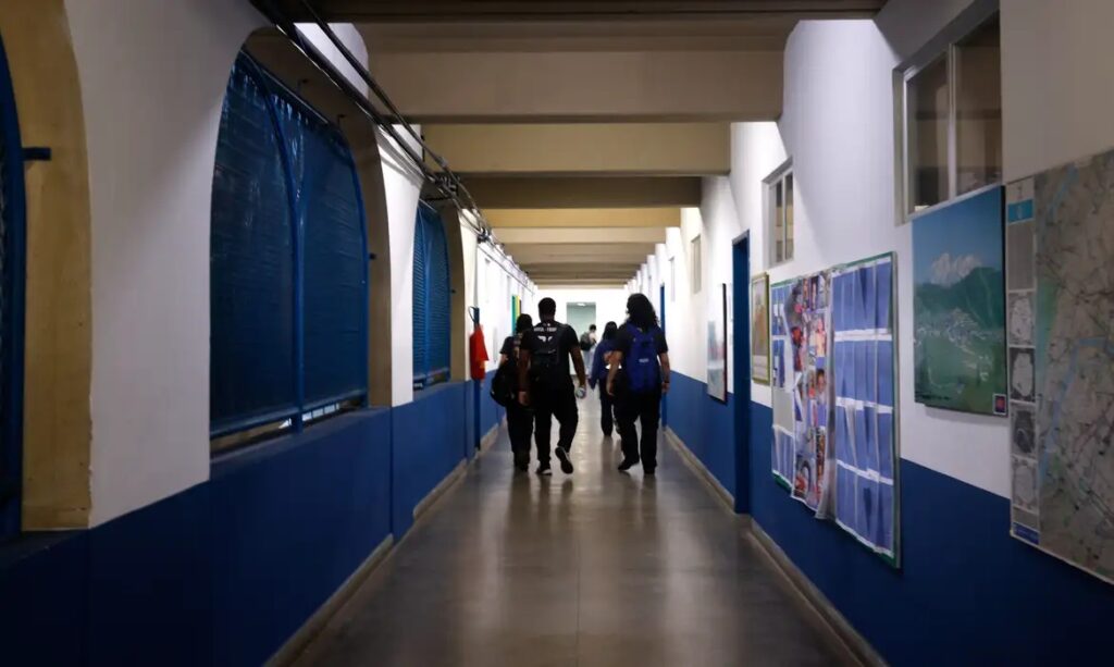 Pessoas caminhando por um corredor escolar com paredes azuis e brancas.