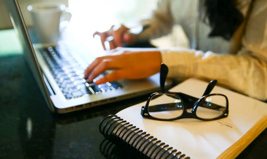 Mãos digitando em um notebook com óculos e caderno em primeiro plano.