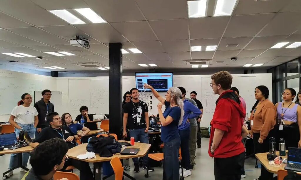 Uma mulher aponta para uma tela com gráficos, cercada por estudantes em sala de aula.