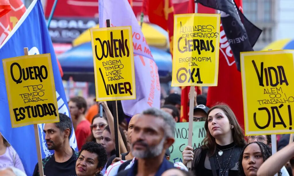 Pessoas em protesto segurando cartazes amarelos com as frases 'Jovens Exigem Tempo Livre Para Viver' e 'Auxiliares de Serviços Gerais Exigem o Fim da 6X1'.