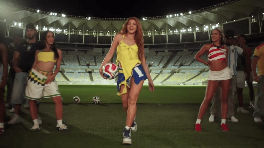 Shakira sorrindo em campo de futebol, vestindo roupa amarela e azul, segurando uma bola.