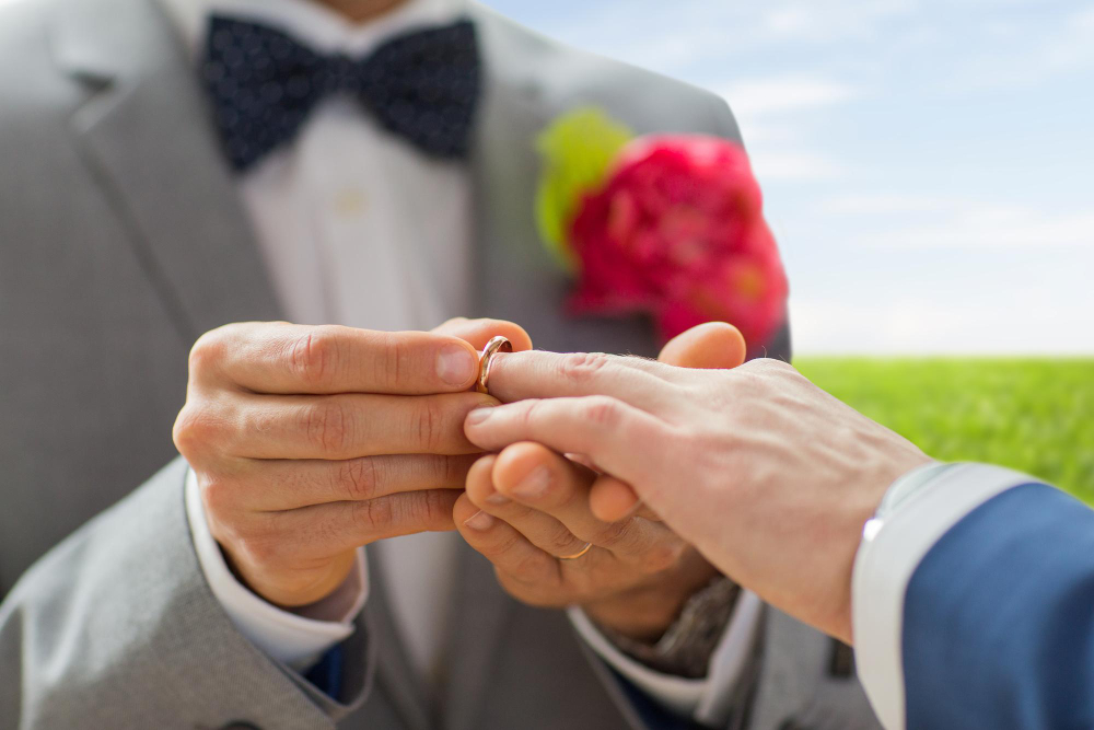 Mãos trocando alianças de casamento em cerimônia ao ar livre.