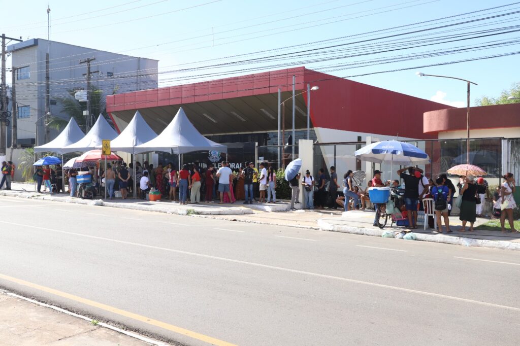Fila de pessoas em frente a um Fórum Eleitoral sob tendas e sol forte.