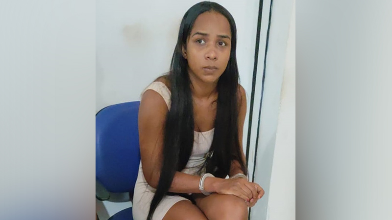 Mulher jovem de cabelo preto liso e vestido claro sentada em cadeira azul, algemada, com expressão séria.