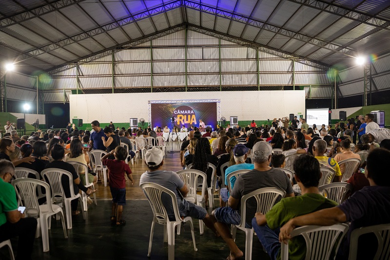 Pessoas sentadas em cadeiras brancas assistindo a um evento em palco com telão "CÂMARA NA RUA".