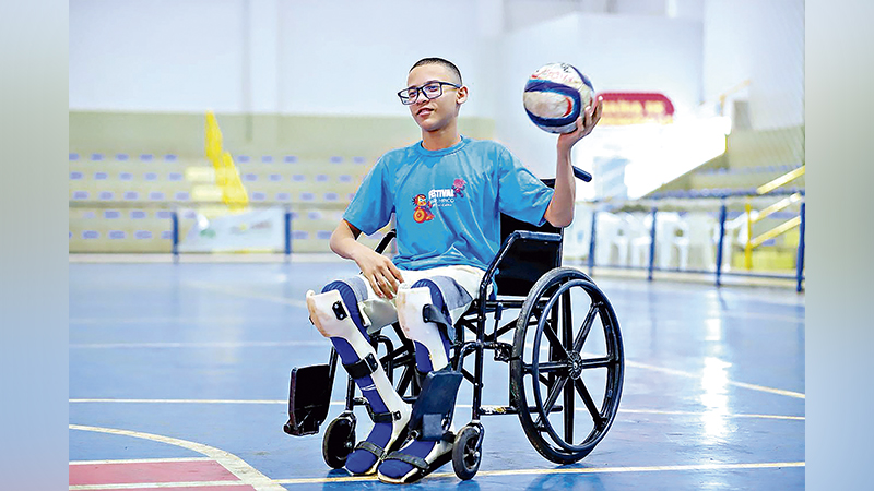 Jovem em cadeira de rodas com órteses nas pernas, segurando uma bola, em quadra esportiva.