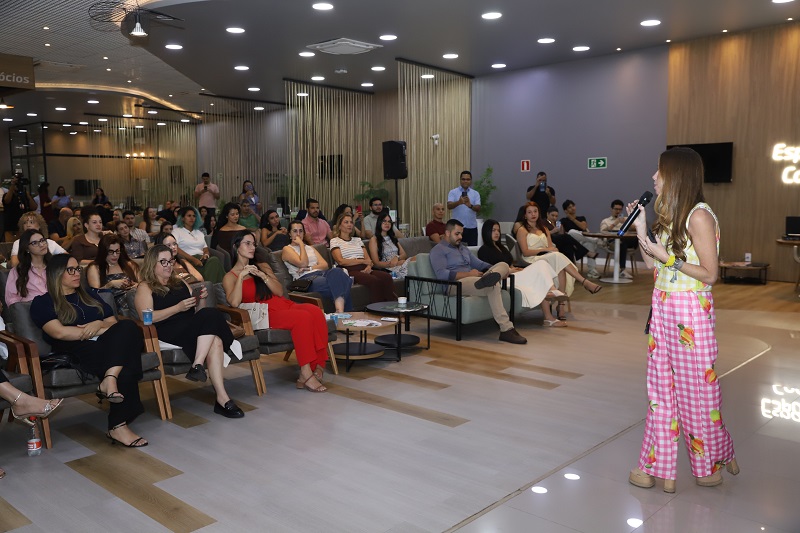 Palestrante mulher em pé, com microfone, interagindo com uma plateia atenta em um auditório moderno.
