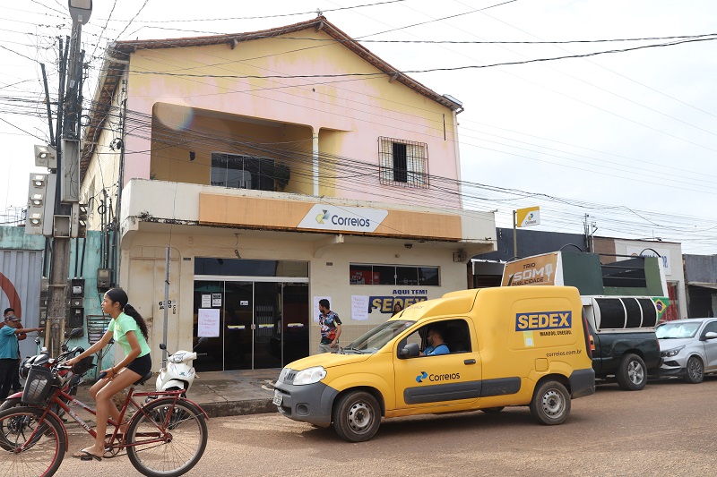 Agência dos Correios com van amarela e jovem de bicicleta na rua.