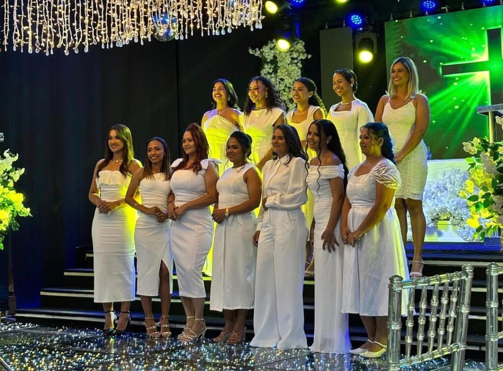 Grupo de mulheres sorrindo em trajes brancos em um palco iluminado.