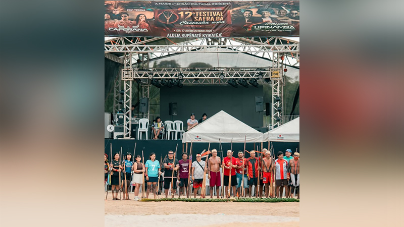 Grupo de indígenas com lanças e bastões em frente a um palco de festival cultural.