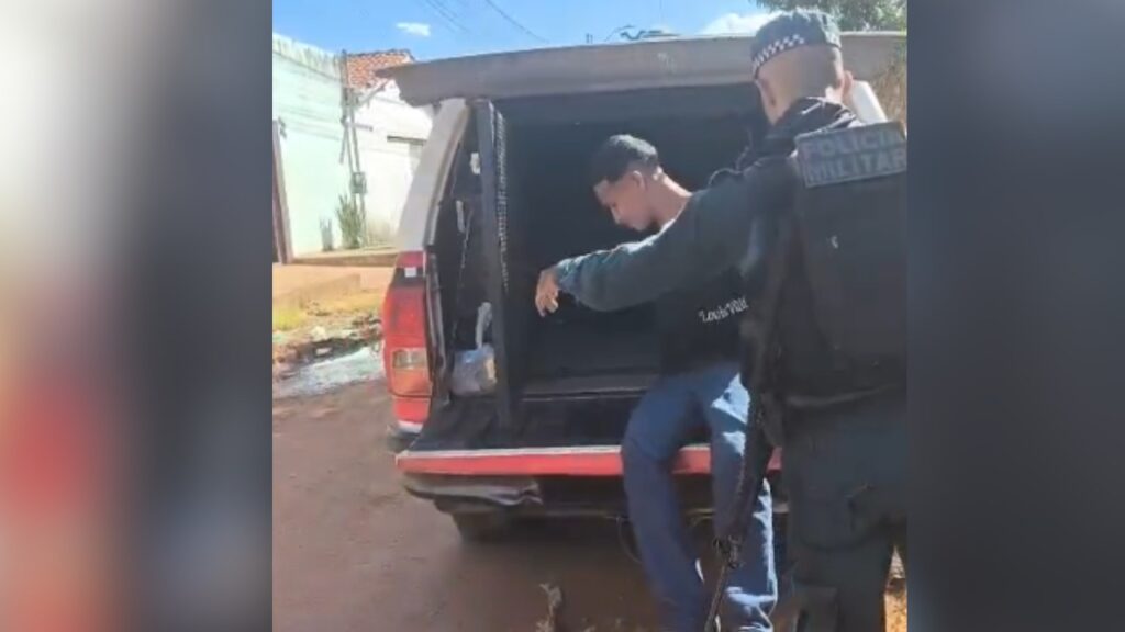 Jovem sendo colocado na caçamba de uma caminhonete por policial militar.