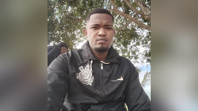 Homem negro vestindo jaqueta preta do Corinthians com fundo de árvores.