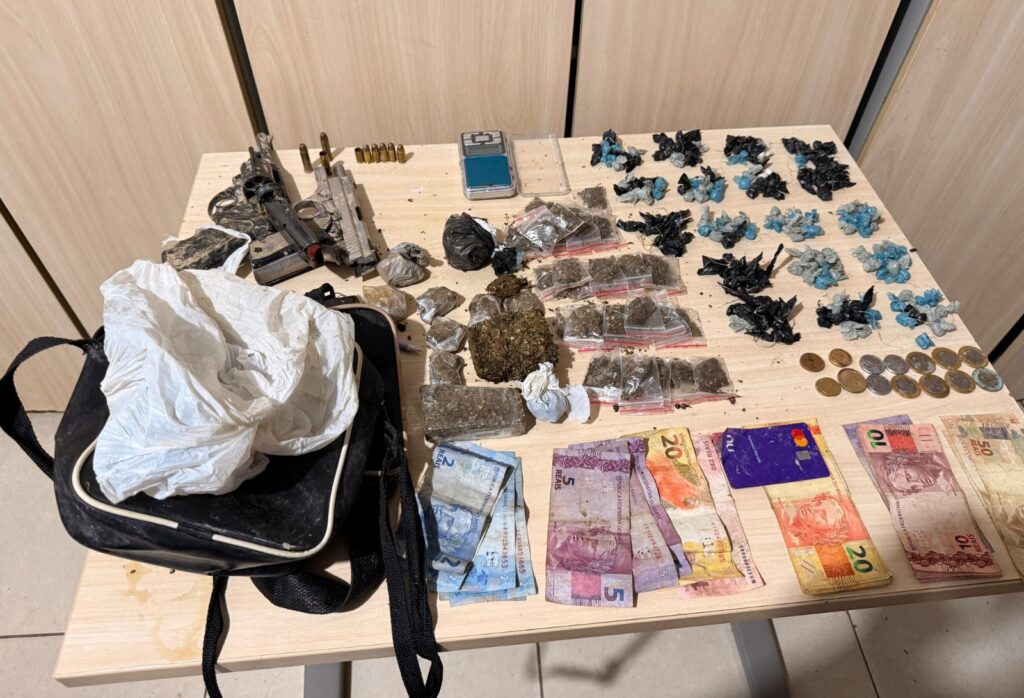 Mesa com arma, munições, drogas (maconha, comprimidos), balança digital e dinheiro em notas.