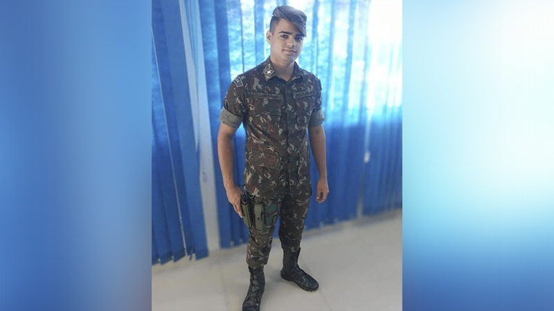 Jovem militar brasileiro em uniforme camuflado, com coldre na perna, em pé em ambiente interno.