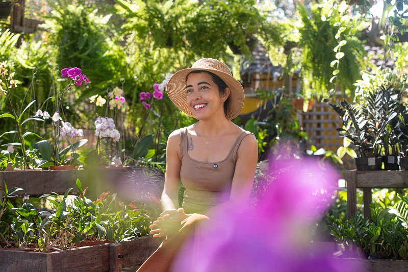 Mulher sorridente com chapéu de palha em um viveiro de plantas com orquídeas coloridas.