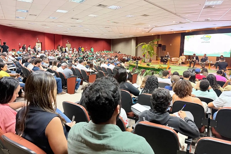 Auditório lotado com pessoas assistindo a um painel de discussão em um palco com tela de apoio.