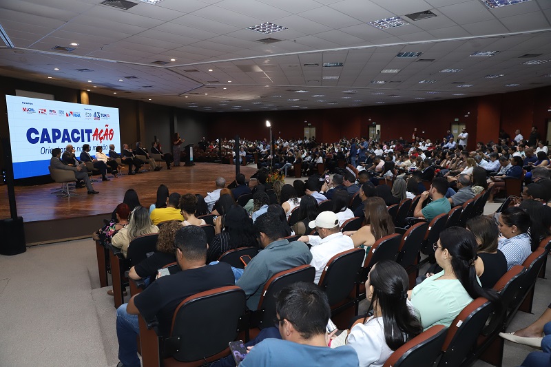 Auditório lotado com pessoas assistindo a um evento de capacitação. Um painel de pessoas está no palco em frente a uma tela grande.