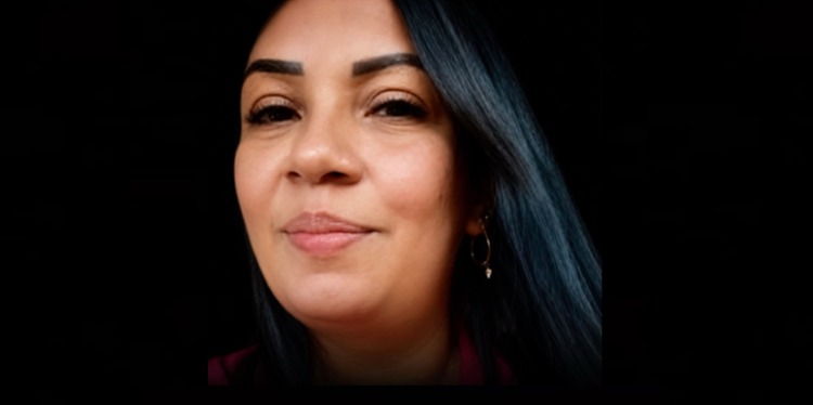 Mulher sorrindo com cabelos escuros e brinco em fundo preto.