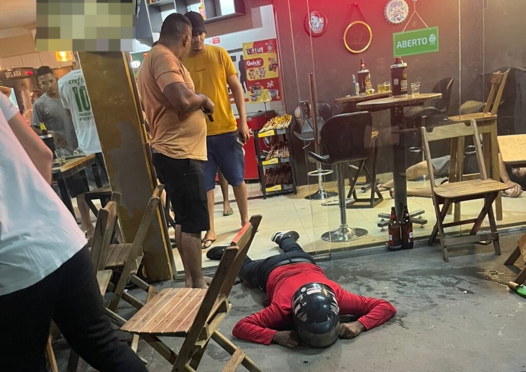 Homem de capacete caído no chão em frente a um bar, com outras pessoas ao redor.