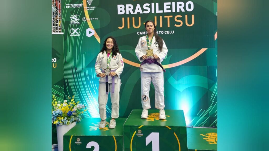 Duas mulheres com quimonos e faixas roxas em pódio de jiu-jitsu, uma com medalha de ouro e outra com prata.