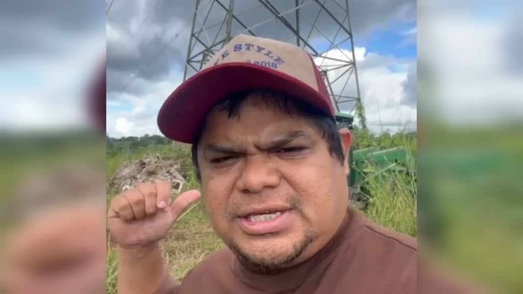 Homem com boné gesticulando em campo aberto com torre de energia.