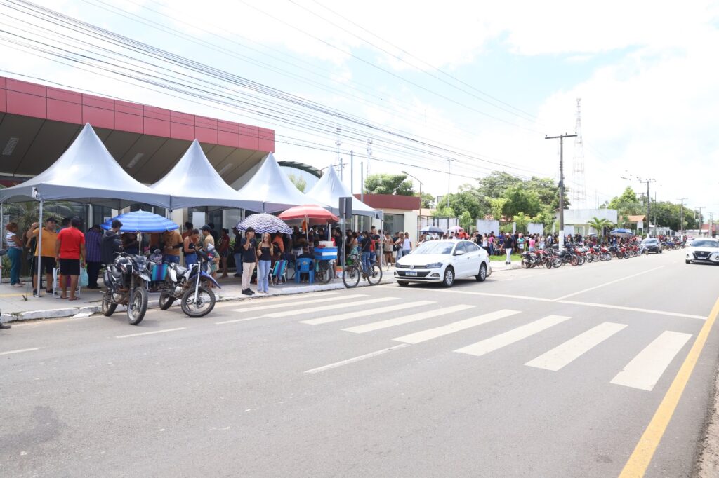 Vista de uma rua movimentada com pessoas sob tendas, motocicletas e um carro branco em uma faixa de pedestres.