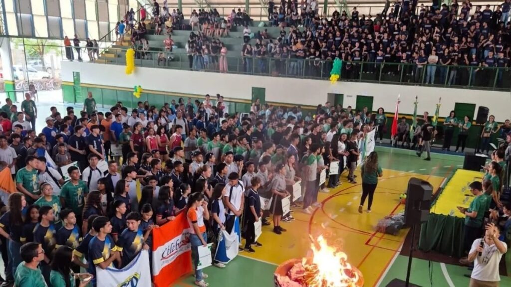 Cerimônia de abertura de evento escolar em ginásio com estudantes e pira olímpica acesa.