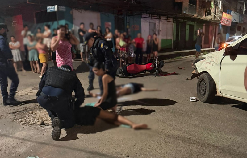 Cena de acidente de trânsito noturno com carro branco e moto vermelha danificados, pessoas caídas na rua e socorristas.