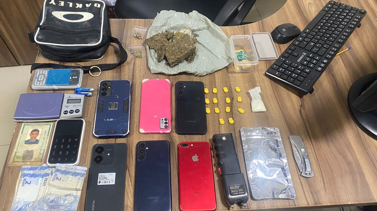 Itens apreendidos em mesa de madeira, incluindo drogas, celulares, balanças de precisão e uma arma de choque.